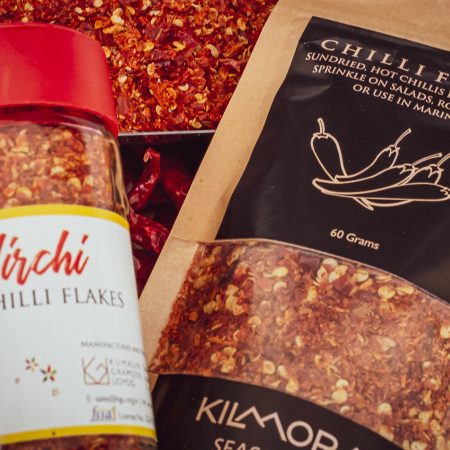 Glas jar with Mirchi written on label. And a pouch with Chilli Flakes written on it. Resting atop a bed of dried red chillies.