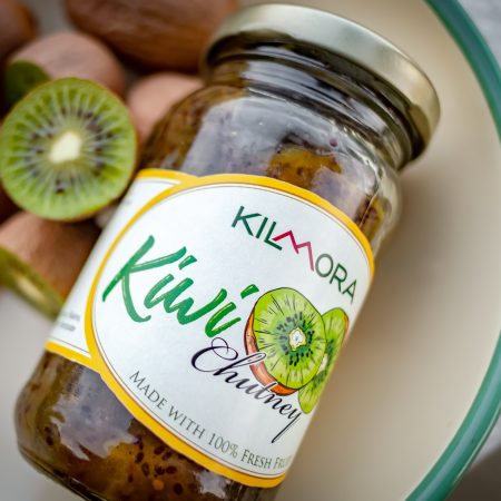 Jar of Kilmora Kiwi Chutney kept next to some ripe kiwi fruits and one half of a kiwi fruit cut