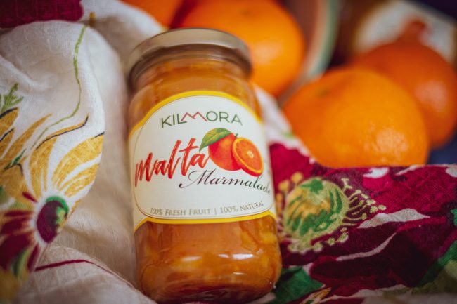 Jar of Malta Marmalade lying atop floral cloth napkin with some Malta fruits in the background