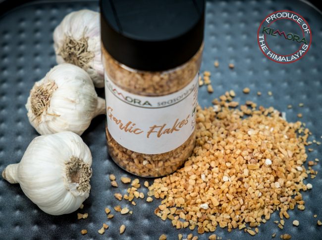 Top shot of a jar of Garlic Flakes, with some garlic flakes sprinkled around it and three bulbs of raw garlic next to the jar