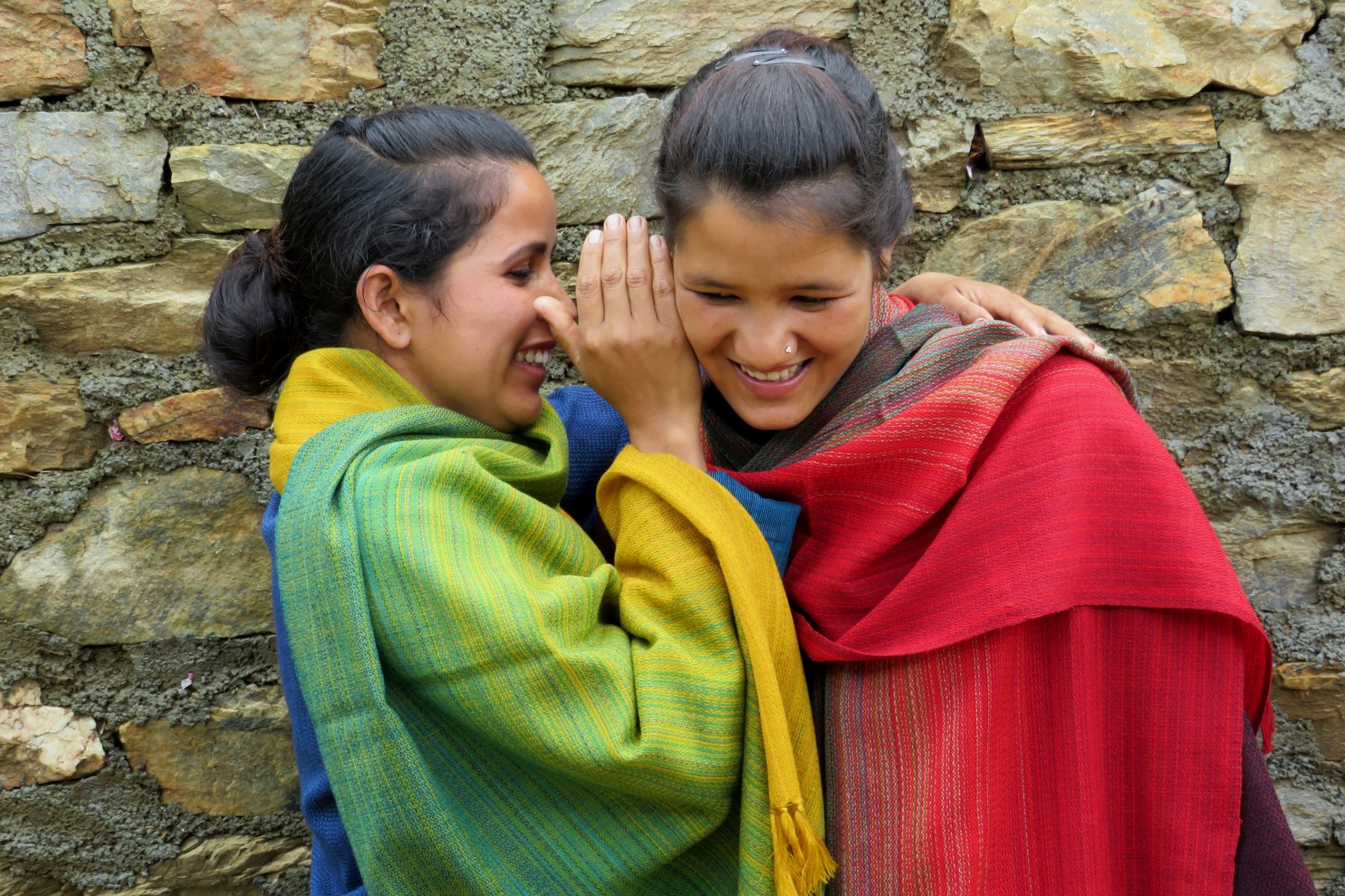 Two women wearing colourful shawls. Woman on the left whispering something into the ear of the woman on the right. They are both smiling.