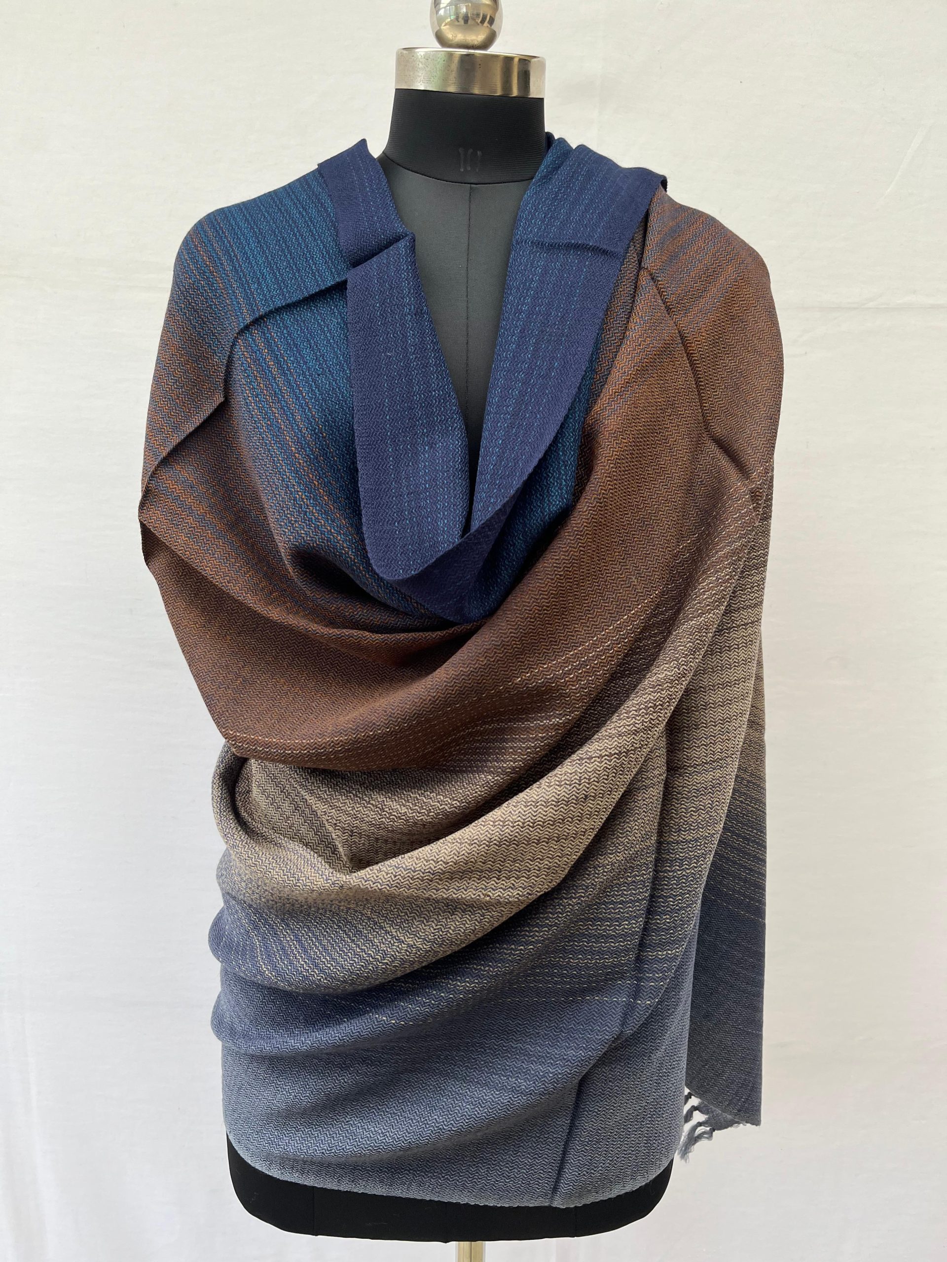 Ombre merino wool shawl in shades of aegean blue with creams and browns in the middle and a deep spruce blue at the other end. Here draped on a mannequin.