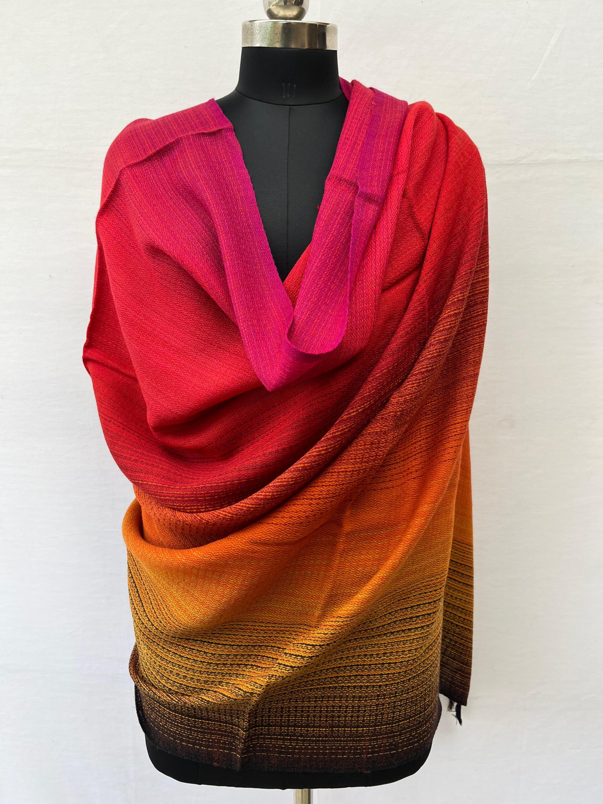 Ombre hand woven merino wool shawl in shades starting with deep red, scarlet, orange and ending with hint of pine green. Draped on a mannequin.