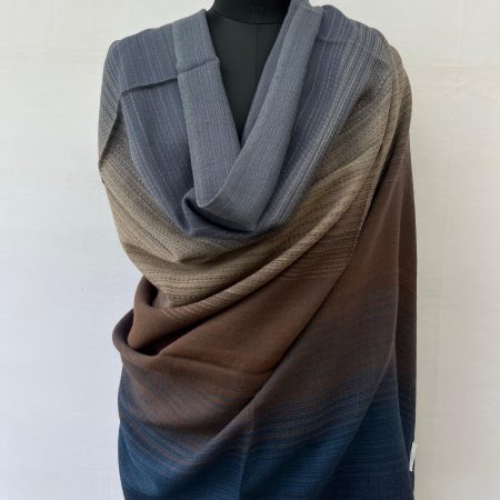 Ombre merino wool shawl in shades of aegean blue with creams and browns in the middle and a deep spruce blue at the other end. Here draped on a mannequin.