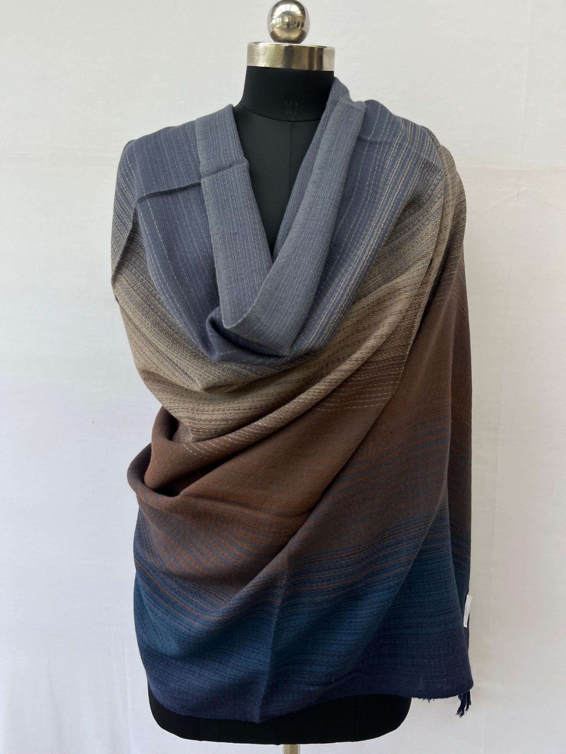 Ombre merino wool shawl in shades of aegean blue with creams and browns in the middle and a deep spruce blue at the other end. Here draped on a mannequin.