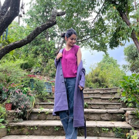 Model is wearing a hand-knit cotton jaali top in magenta. Paired with a pair of jeans and a long coat