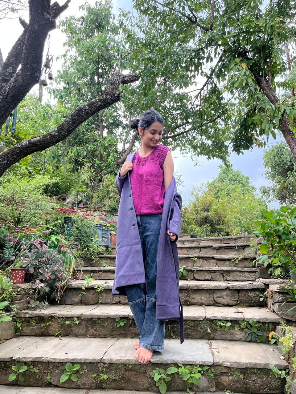Model is wearing a hand-knit cotton jaali top in magenta. Paired with a pair of jeans and a long coat