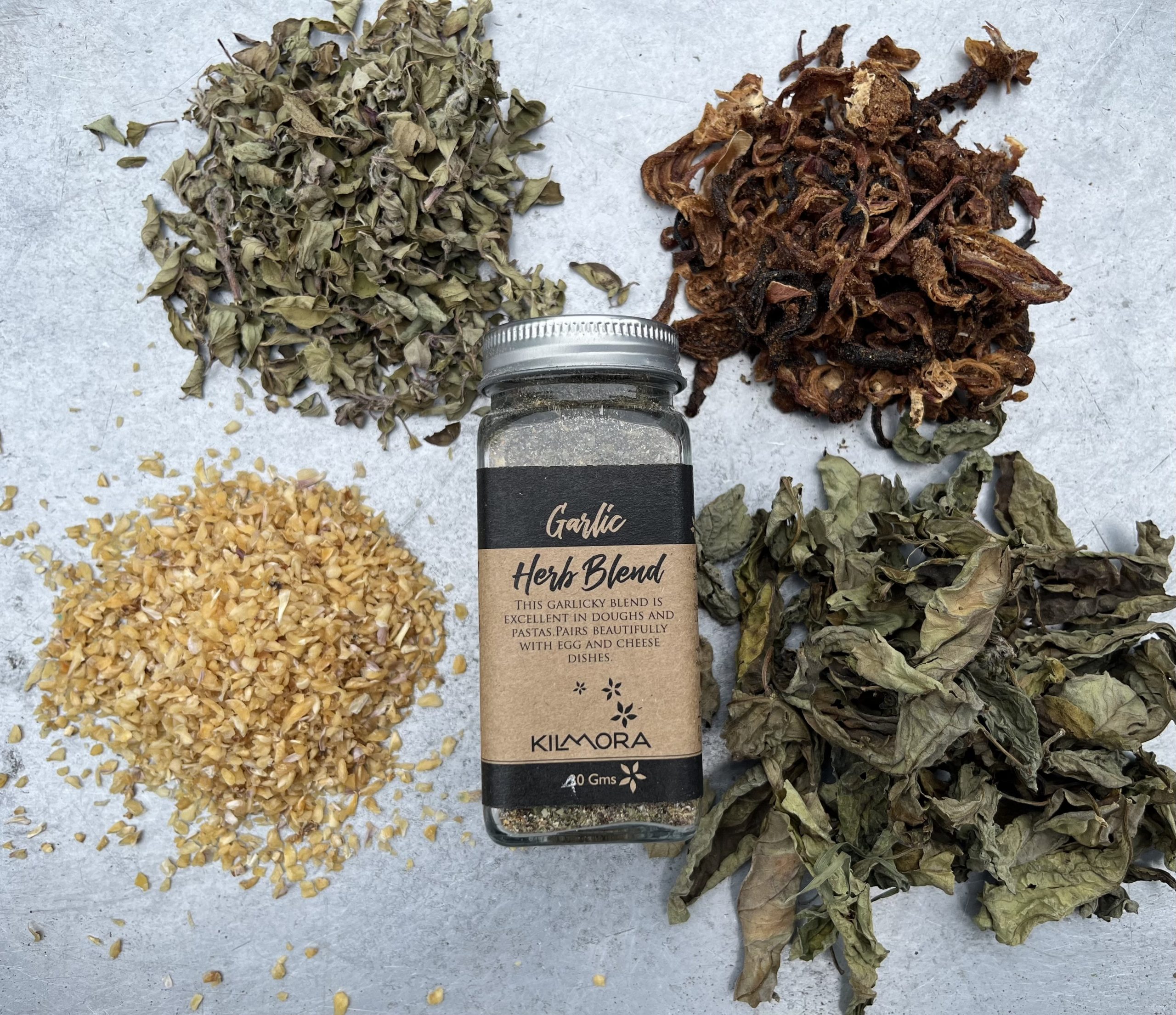 A flat lay photograph of a jar in the center with a Kraft paper label saying Garlic Herb Blend. With sun-dried garlic flakes, oregano, dried onion slices and basil placed in a circle around it.