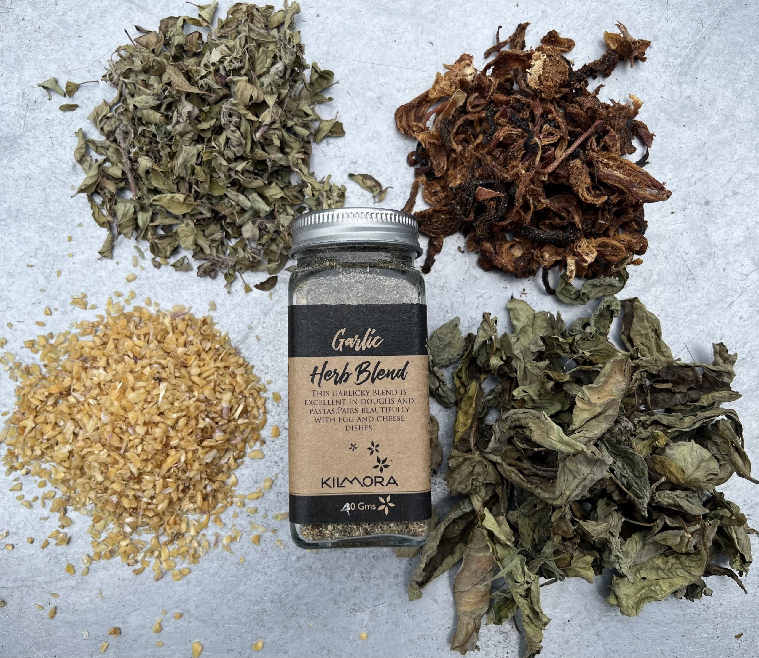 A flat lay photograph of a jar in the center with a Kraft paper label saying Garlic Herb Blend. With sun-dried garlic flakes, oregano, dried onion slices and basil placed in a circle around it.