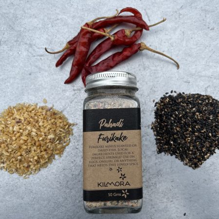 A flat lay photograph of a jar in the center with a Kraft paper label saying Pahadi Furikake. With dried red chillies, sun-dried garlic flakes and sesame seeds placed in a circle around it.