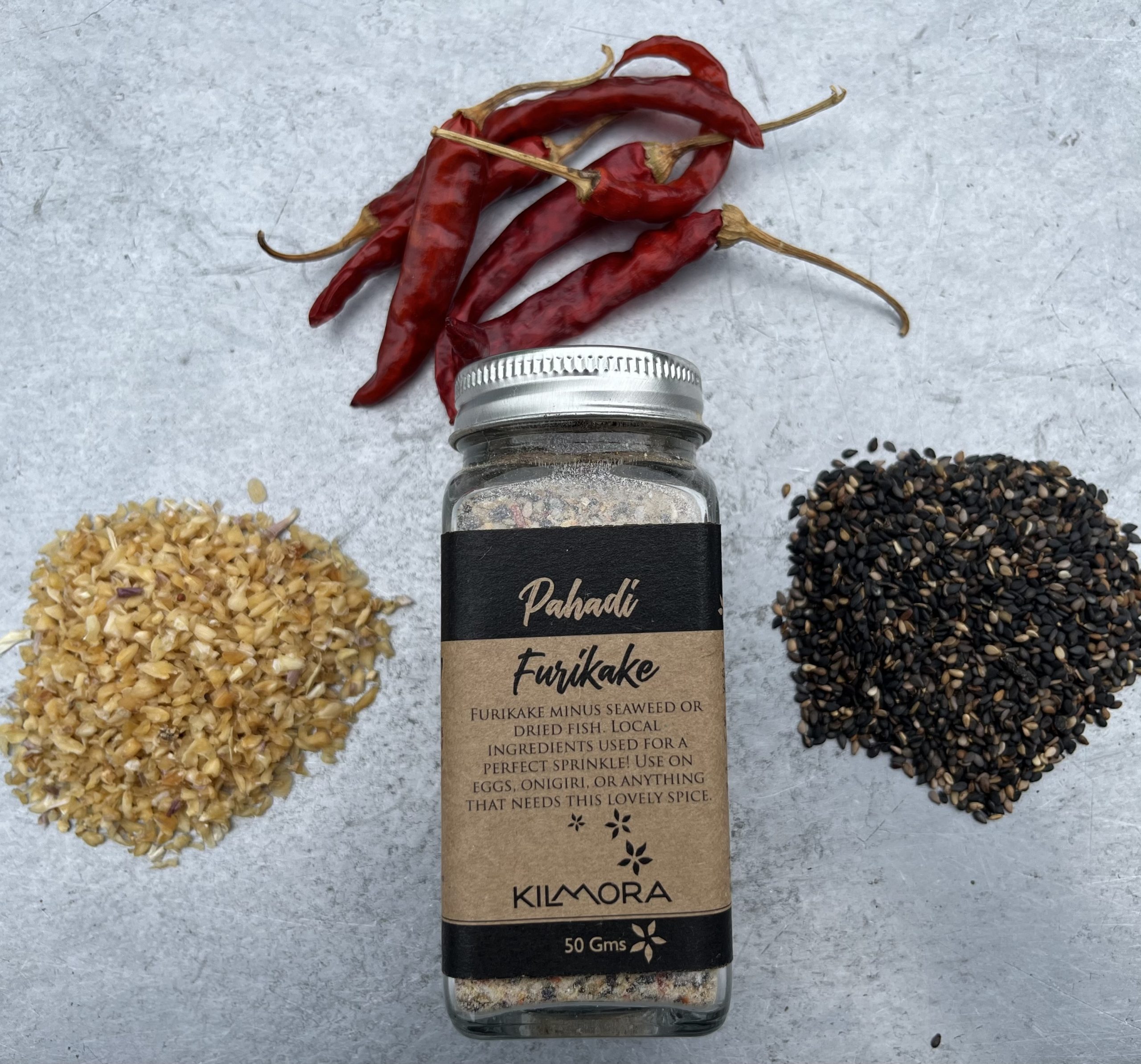 A flat lay photograph of a jar in the center with a Kraft paper label saying Pahadi Furikake. With dried red chillies, sun-dried garlic flakes and sesame seeds placed in a circle around it.