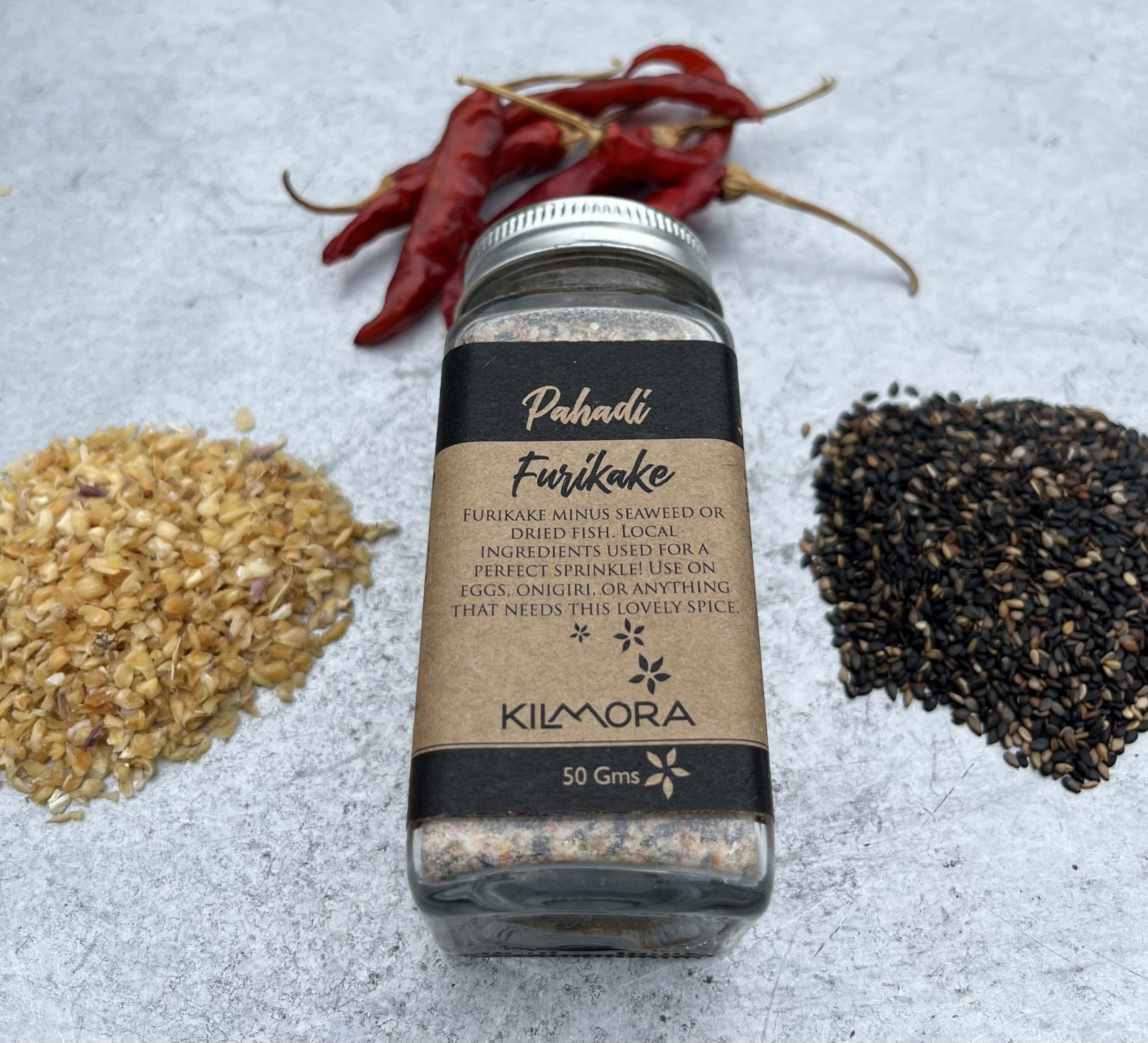 A flat lay photograph of a jar in the center with a Kraft paper label saying Pahadi Furikake. With dried red chillies, sun-dried garlic flakes and sesame seeds placed in a circle around it.