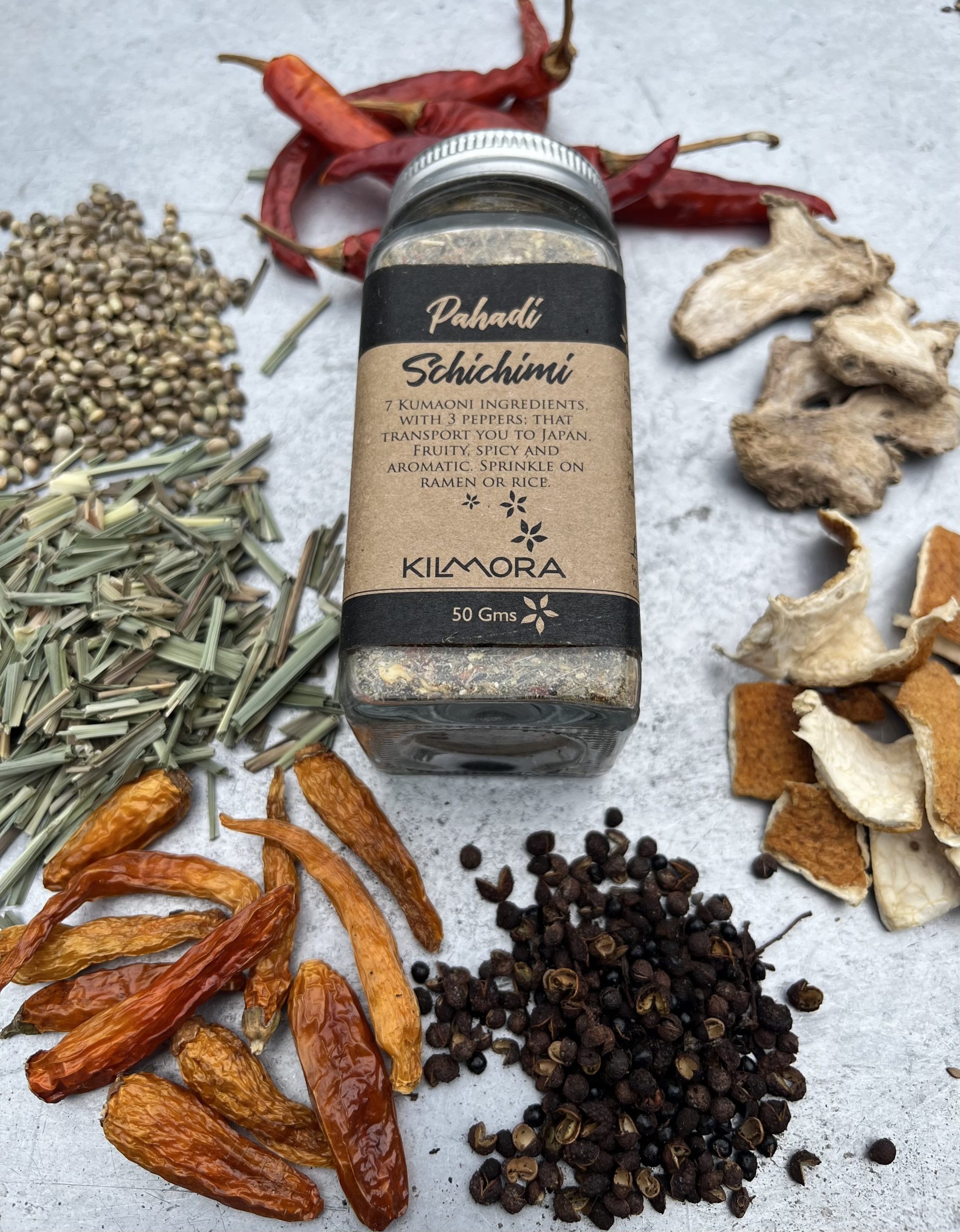 A flat lay photograph of a jar in the center with a Kraft paper label saying Pahadi Schichimi. With dried red chillies, yellow chillies, citrus peel, timur, dry ginger slices, hemp seeds, lemongrass placed in a circle around it.