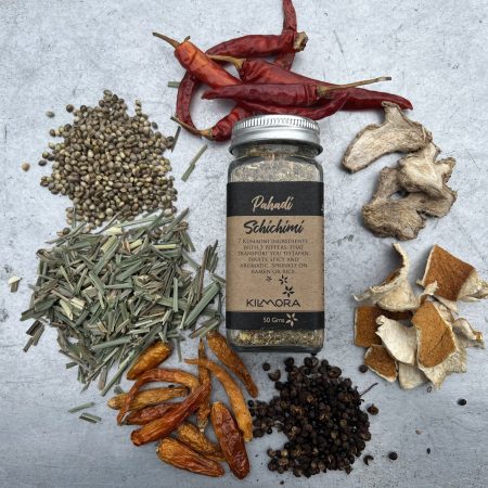 A flat lay photograph of a jar in the center with a Kraft paper label saying Pahadi Schichimi. With dried red chillies, yellow chillies, citrus peel, timur, dry ginger slices, hemp seeds, lemongrass placed in a circle around it.