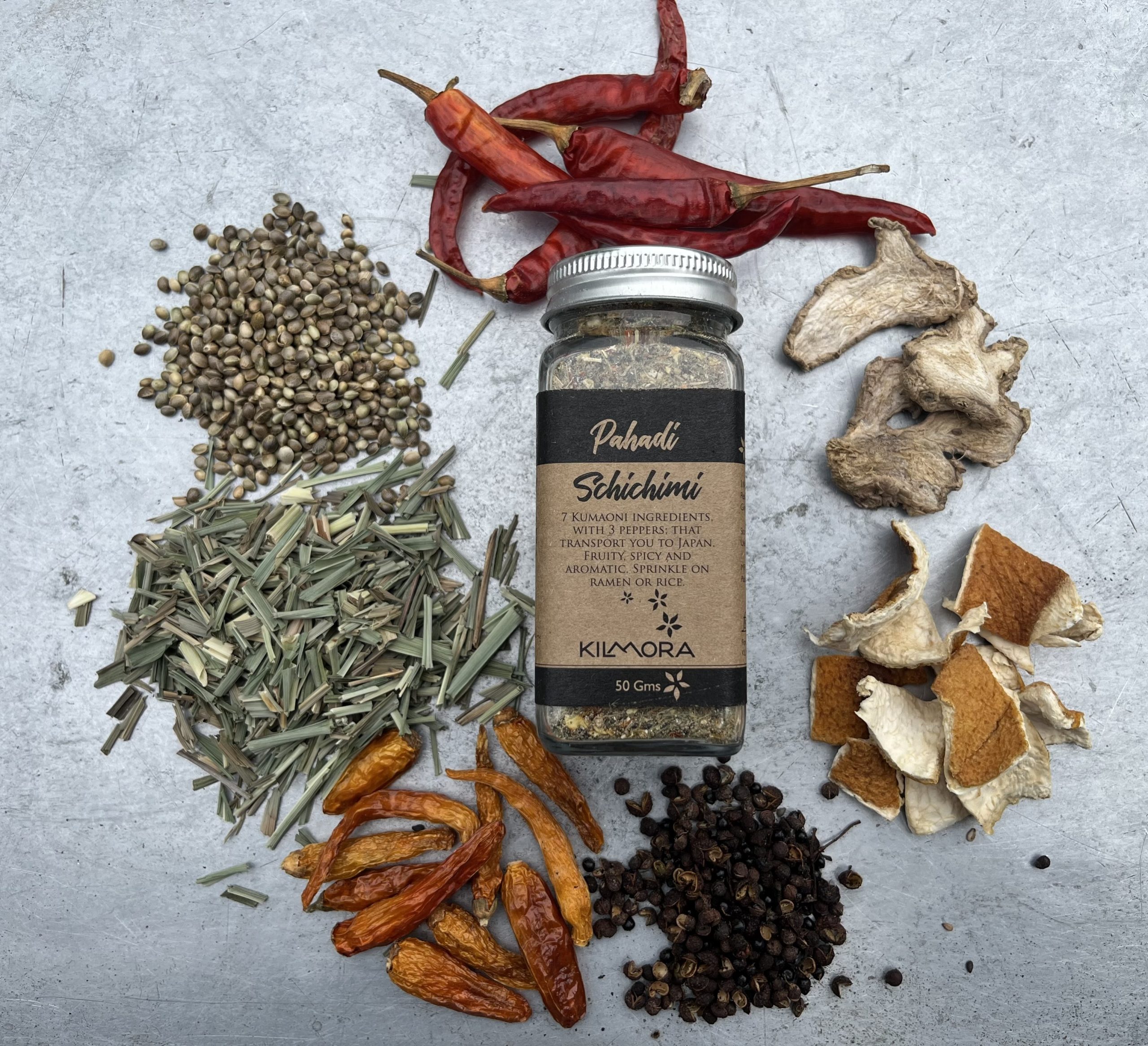 A flat lay photograph of a jar in the center with a Kraft paper label saying Pahadi Schichimi. With dried red chillies, yellow chillies, citrus peel, timur, dry ginger slices, hemp seeds, lemongrass placed in a circle around it.