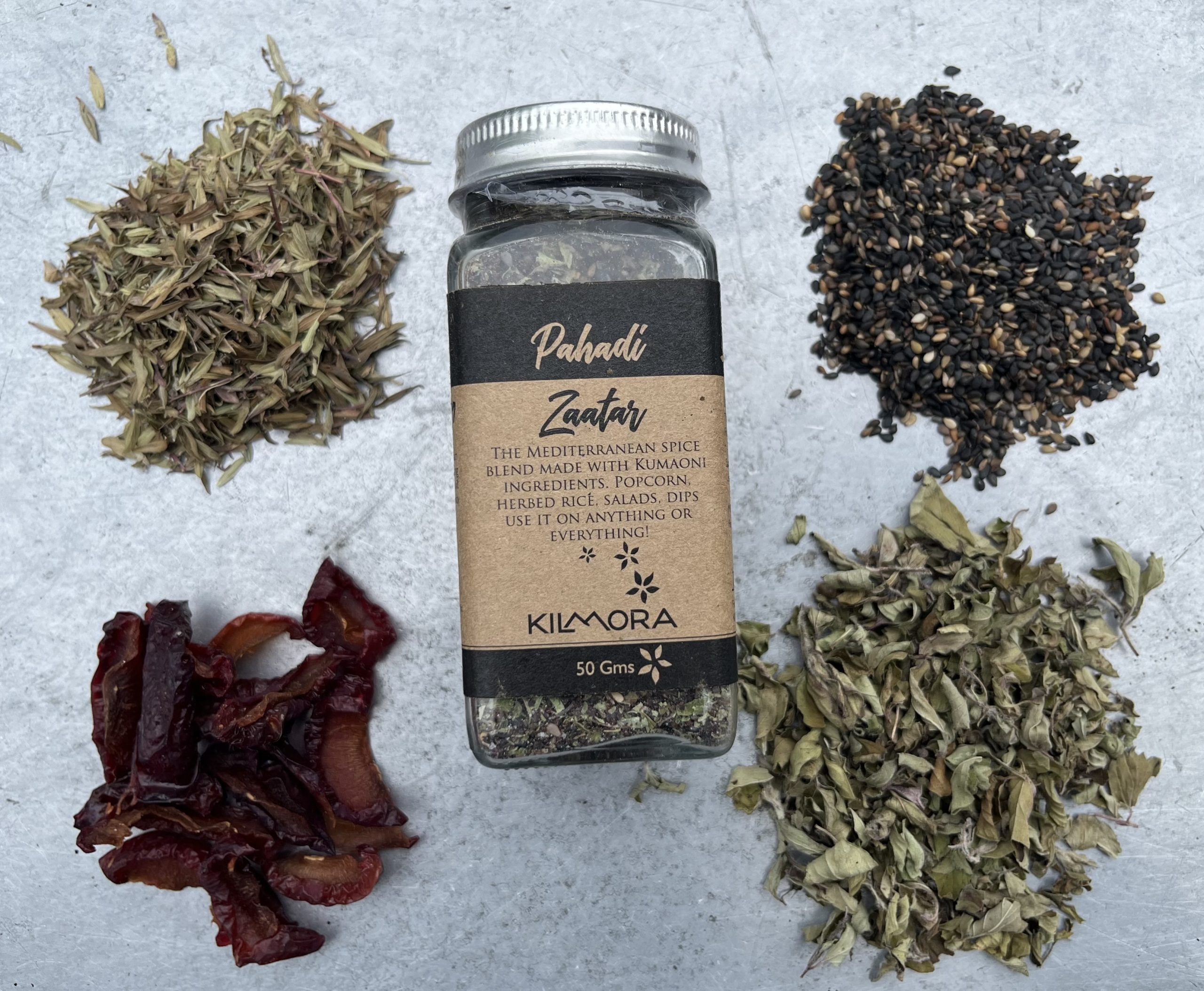A flat lay photograph of a jar in the center with a Kraft paper label saying Pahadi Zaatar. With dried plum slices, thyme, oregano and sesame seeds placed in a circle around it.