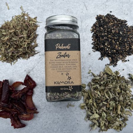 A flat lay photograph of a jar in the center with a Kraft paper label saying Pahadi Zaatar. With dried plum slices, thyme, oregano and sesame seeds placed in a circle around it.