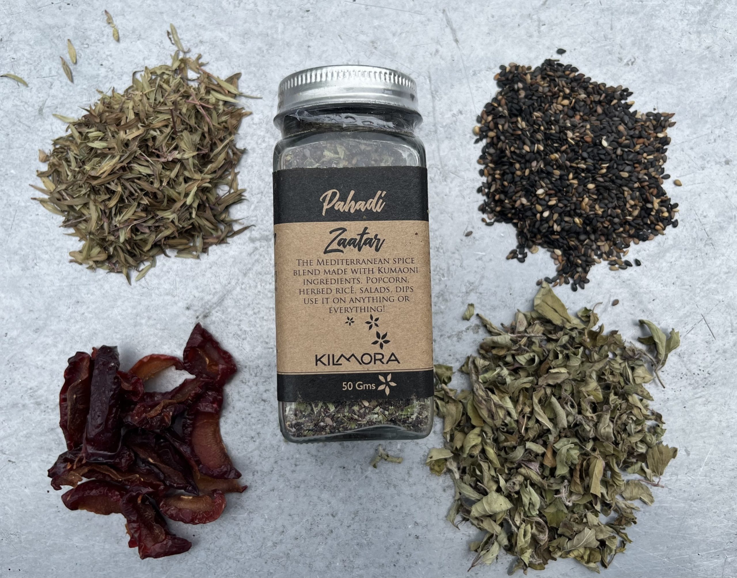 A flat lay photograph of a jar in the center with a Kraft paper label saying Pahadi Zaatar. With dried plum slices, thyme, oregano and sesame seeds placed in a circle around it.