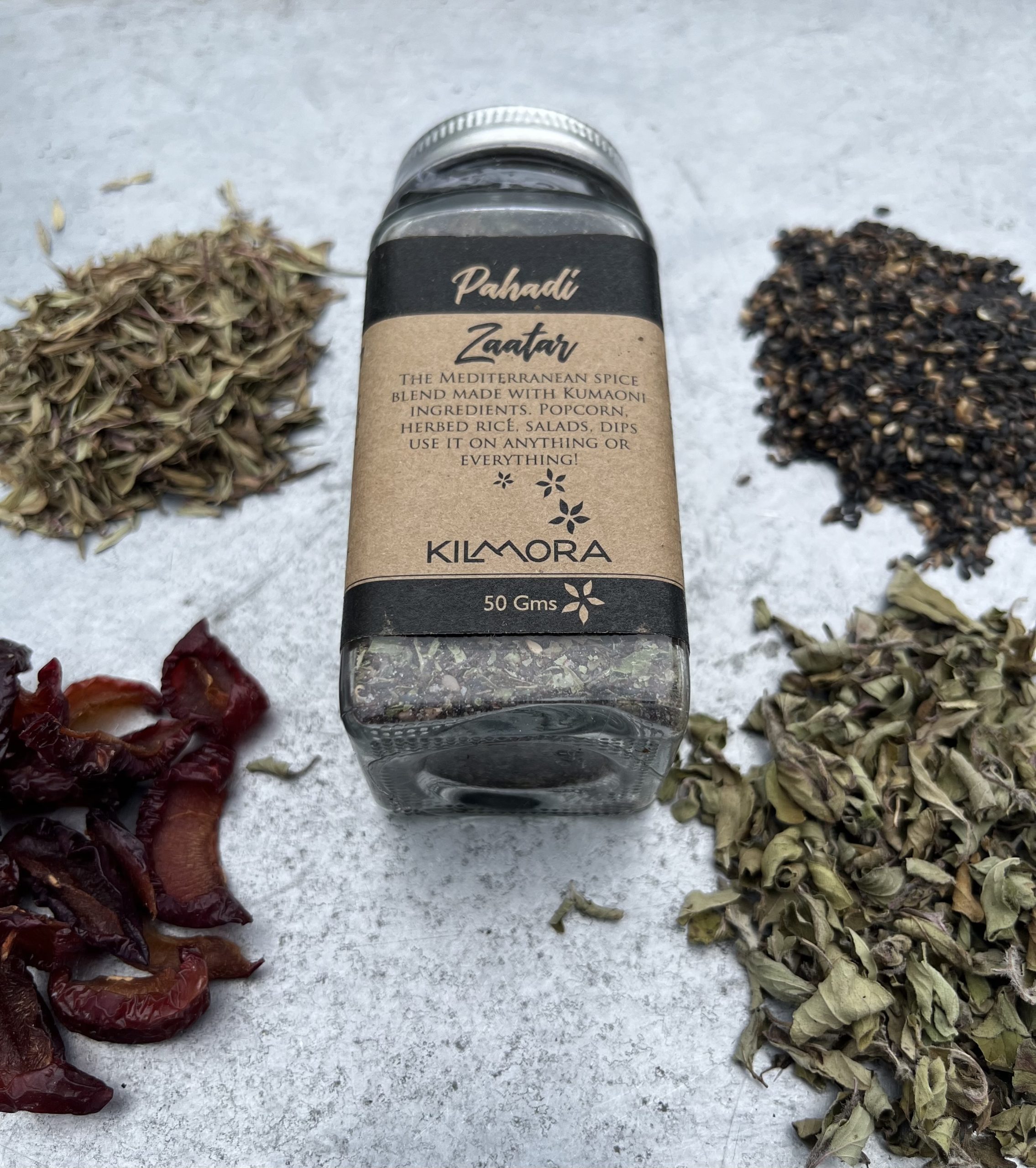 A flat lay photograph of a jar in the center with a Kraft paper label saying Pahadi Zaatar. With dried plum slices, thyme, oregano and sesame seeds placed in a circle around it.