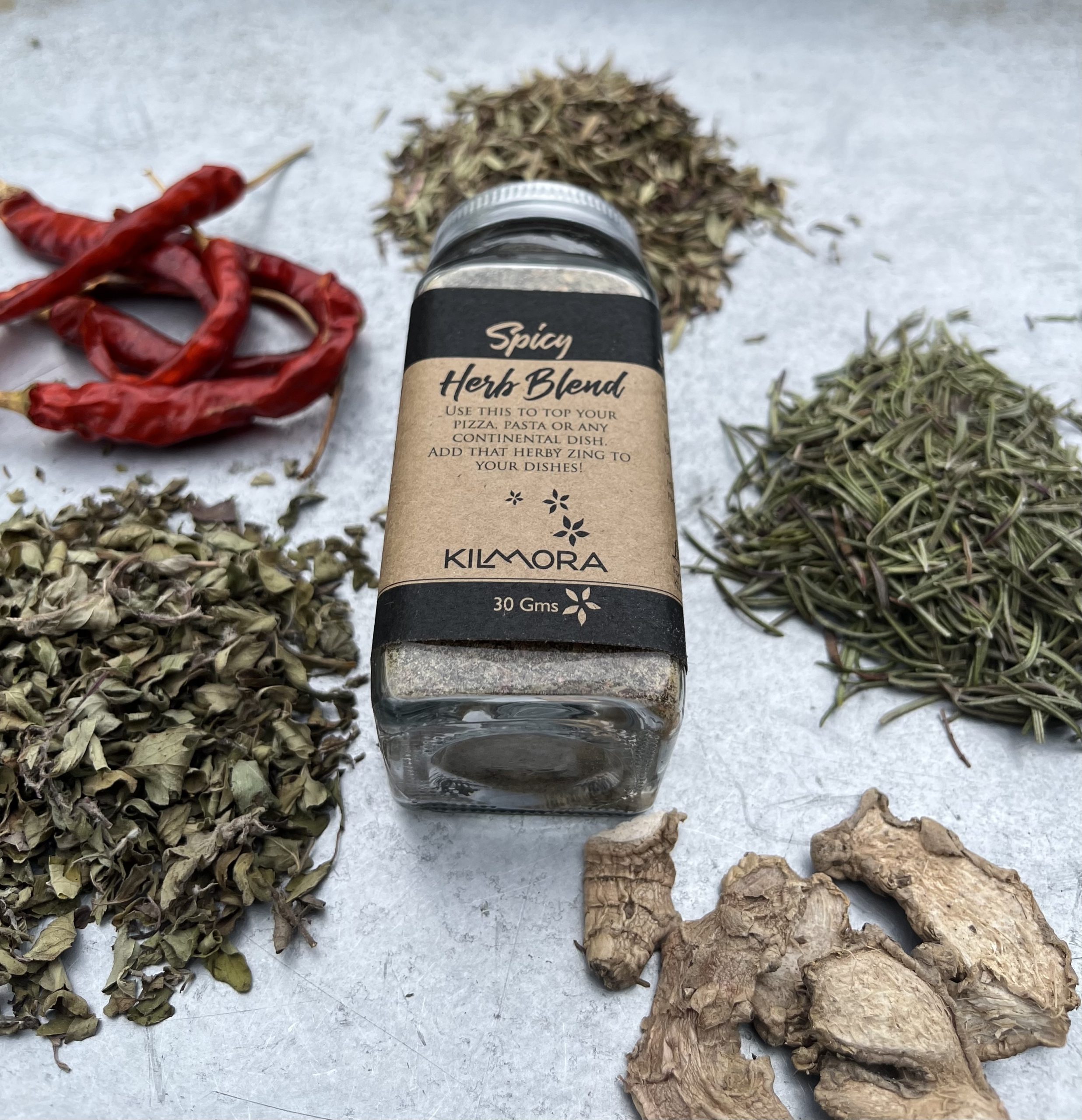 A flat lay photograph of a jar in the center with a Kraft paper label saying Spicy Herb Blend. With dry red chillies, oregano, dry ginger slices and thyme placed in a circle around it.