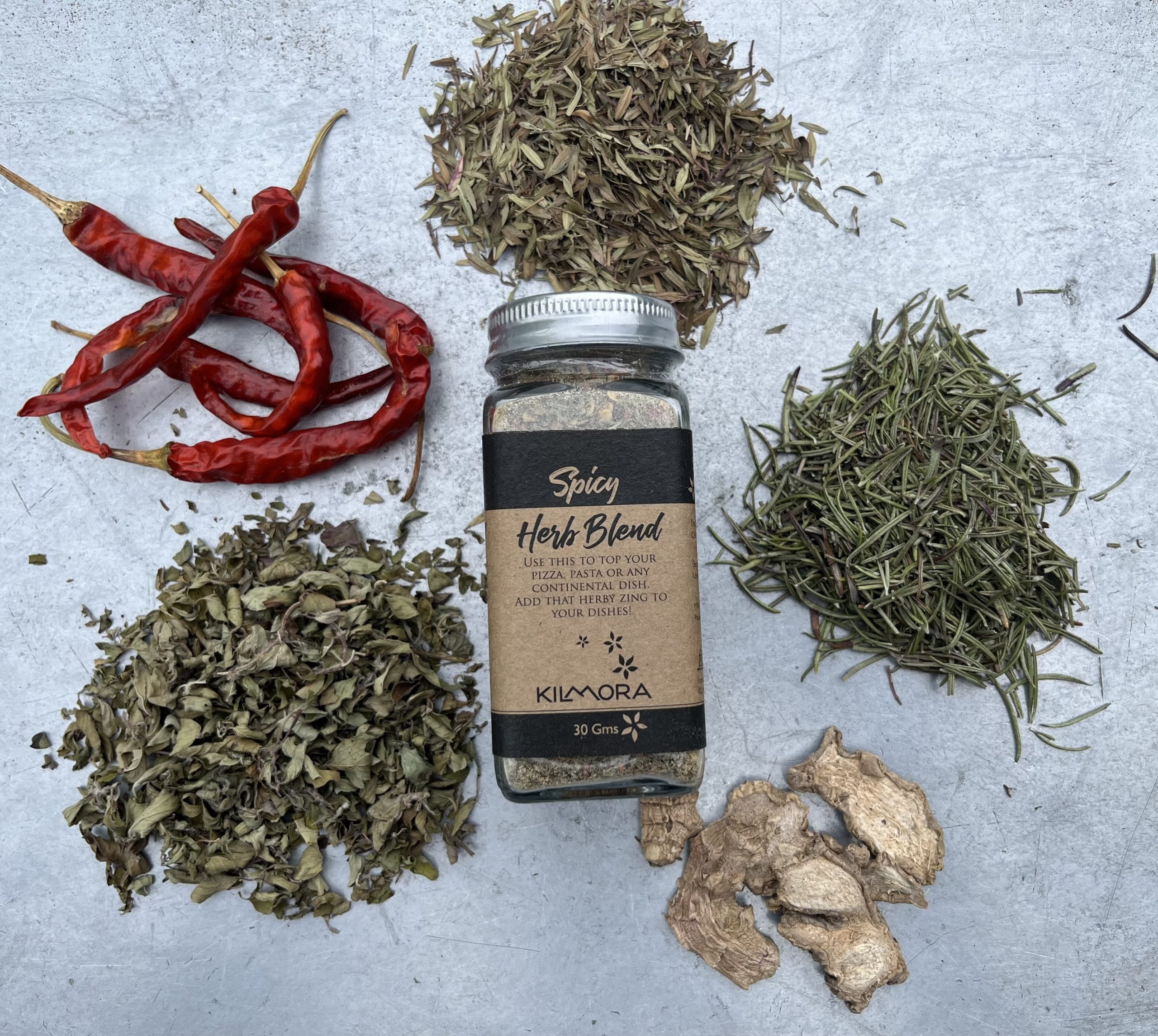 A flat lay photograph of a jar in the center with a Kraft paper label saying Spicy Herb Blend. With dry red chillies, oregano, dry ginger slices and thyme placed in a circle around it.