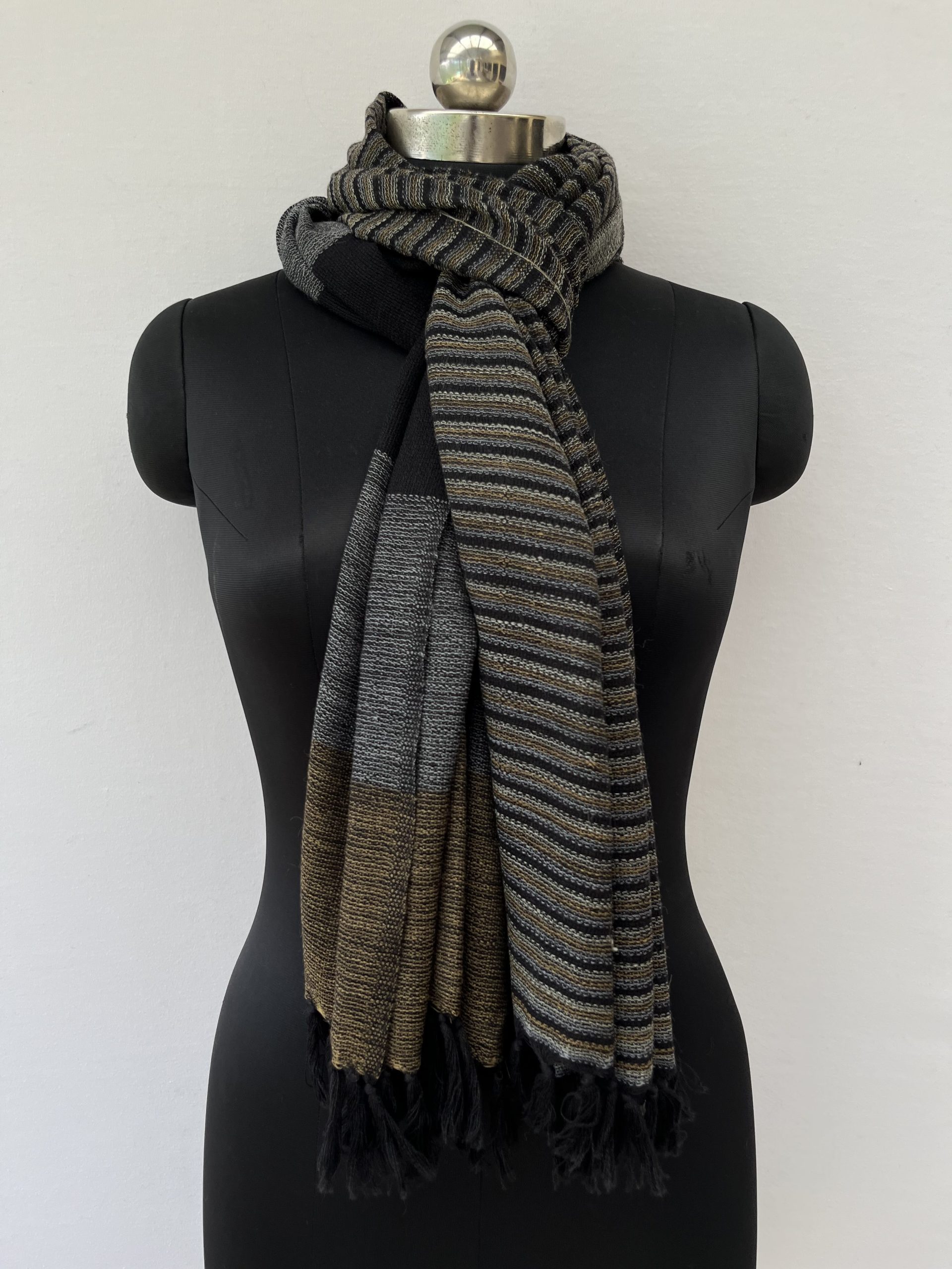 Hand-woven merino wool stole with two different halves to the stole. One half has thin vertical stripes in mustard, silver gray and black. The other half with large vertical stripes of mustard, silver gray and black.